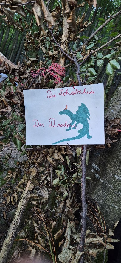 Handgemaltes Schild mit türkisem Drachen und der Aufschrift Die Schatzkiste des Drachen, an einem Ast im Wald befestigt.