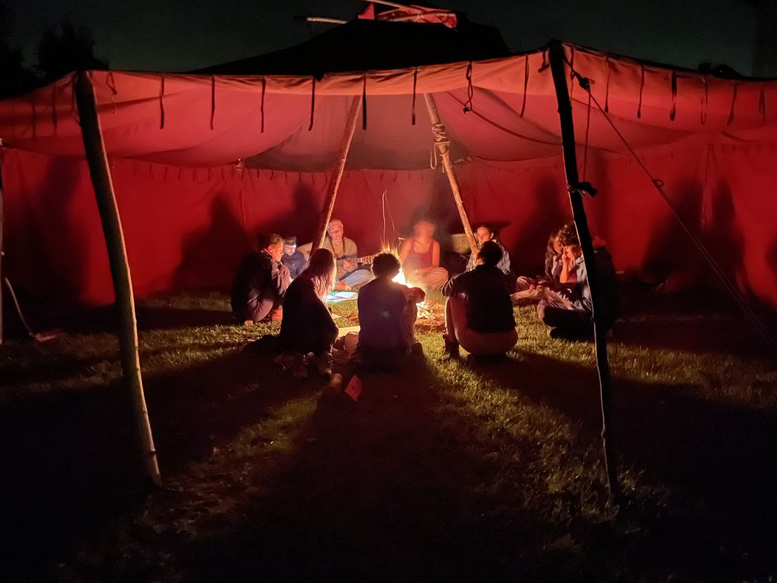 Eine große rote Jurte bei Nacht, innen sitzt eine Gruppe um ein Lagerfeuer.